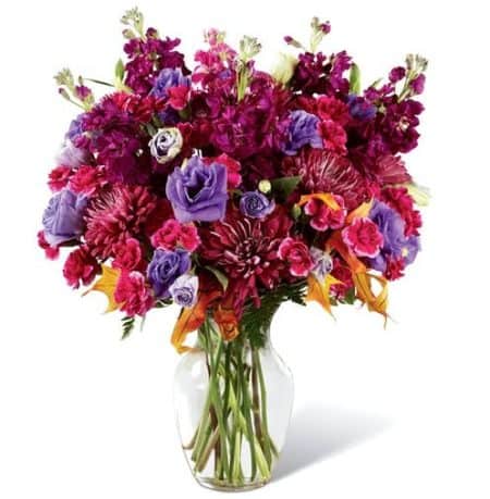 Starting with deep plum chrysanthemums as the base, this arrangement is highlighted by purple gilly flower, purple double lisianthus, hot pink mini carnations, lush green accents, and sunny glycerized oak leaf stems. Presented in a classic clear glass vase
