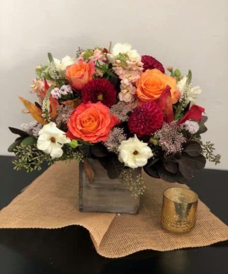 An Assortment of orange roses, burgundy dahlias, peach stock with fillers and fall greens in a wood box.